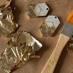 Two earrings mid-process being hand-finished with gold leaf, surrounded by foil and a brush.